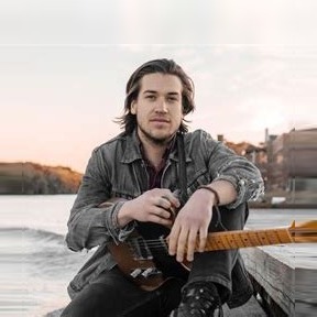 Jackson Stokes sitting on a riverside dock with his electric guitar at sunset